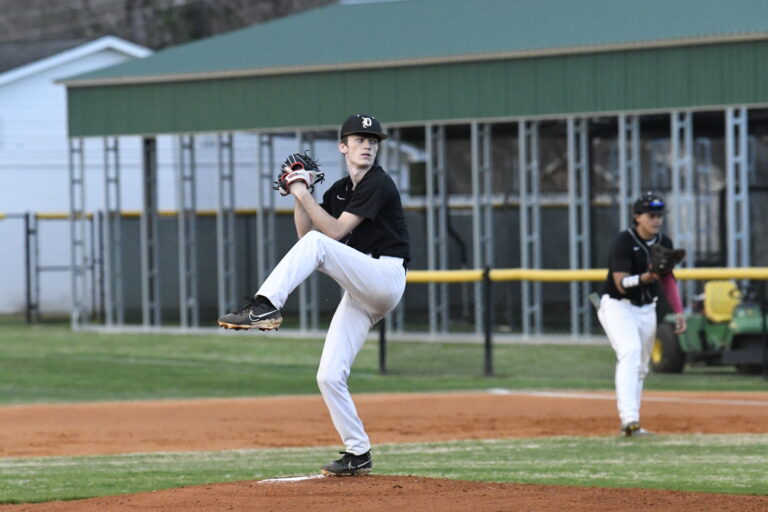 Baseball pitcher in black uniform preparing to throw the ball on the mound, with a teammate in the background near the field.
