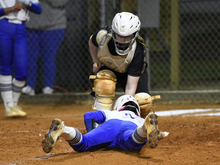 A softball catcher tags a sliding runner at home plate during a game, with dirt flying and intense action evident.