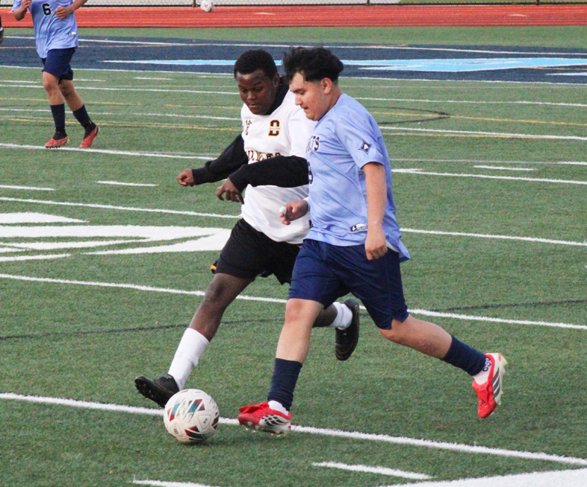 Two soccer players, one in white and one in blue, compete for the ball on a green field during a match.