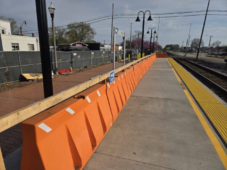 Orange barricades line a train station platform under clear skies, with construction fencing and equipment visible in the background.