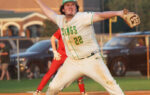 Baseball player in a white and green uniform pitching on a field, with another player in red standing behind and vehicles in the background.