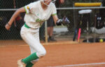A softball player in white and green uniform runs on a dirt field during a game, wearing a helmet and gloves.