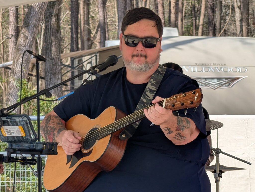 A man wearing sunglasses plays an acoustic guitar outdoors, seated in front of a microphone with trees and musical equipment in the background.