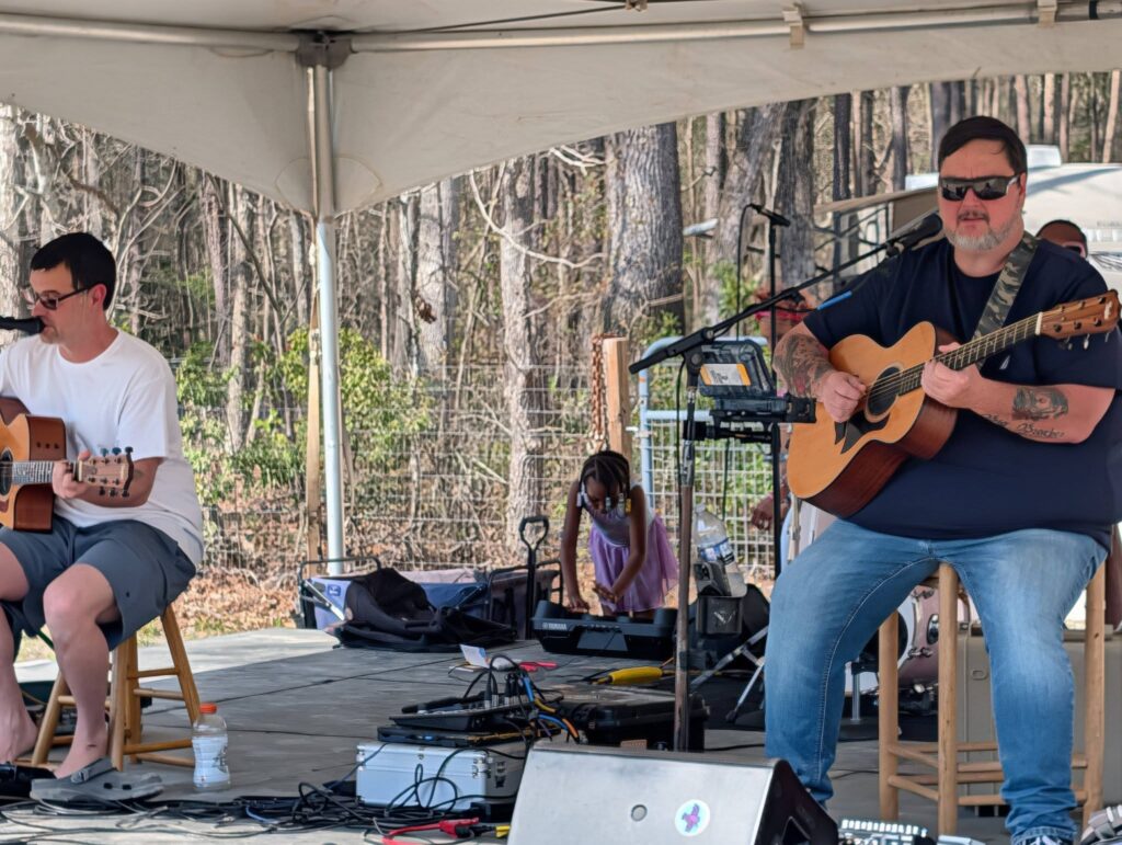 Two musicians performing on an outdoor stage under a tent, one singing and one playing acoustic guitar, with equipment and trees visible in the background.