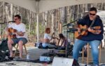 Two musicians play acoustic guitars on an outdoor stage under a tent, while two people sit on the ground behind them surrounded by trees.