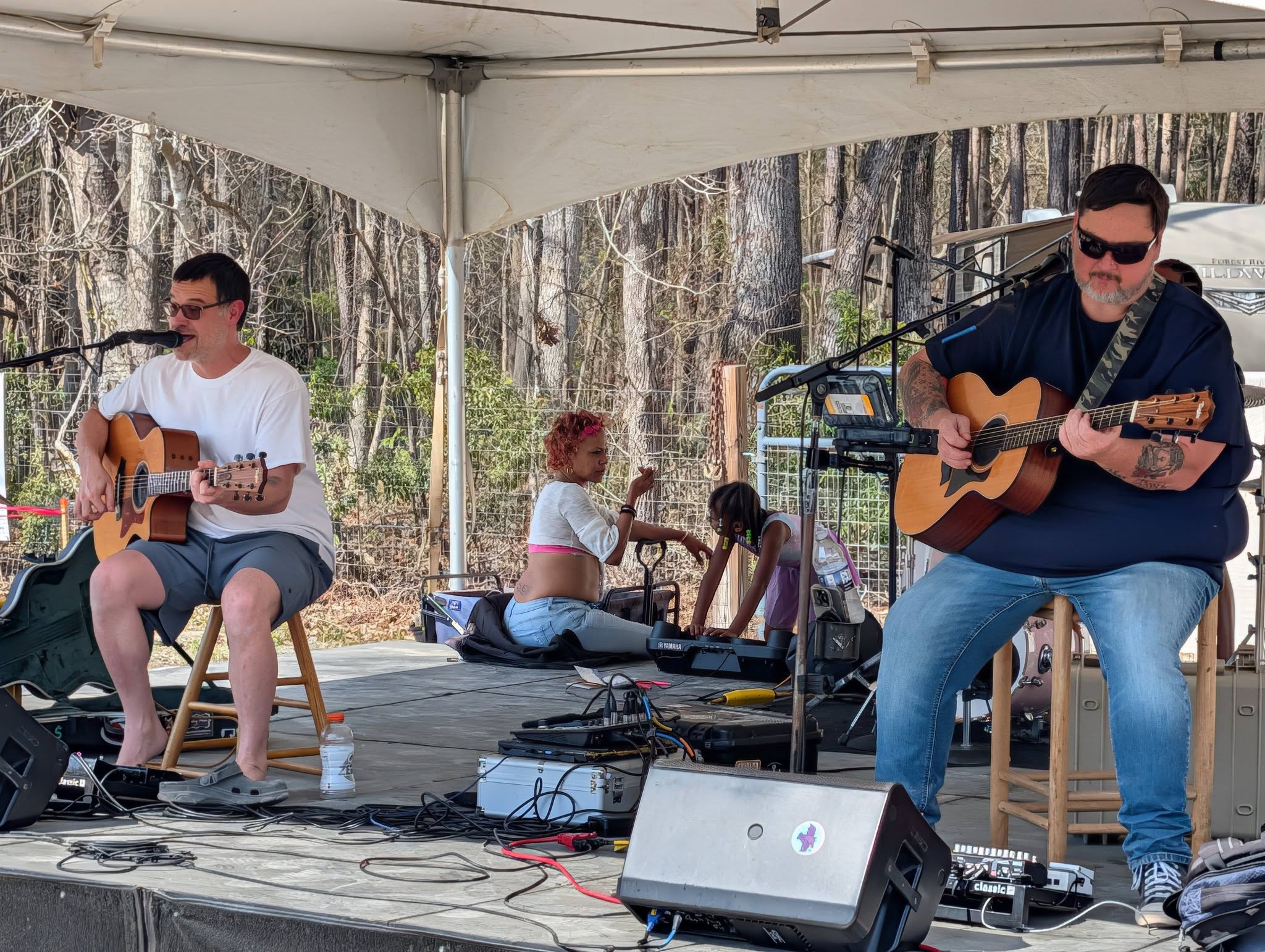 Two musicians play acoustic guitars on an outdoor stage under a tent, while two people sit on the ground behind them surrounded by trees.