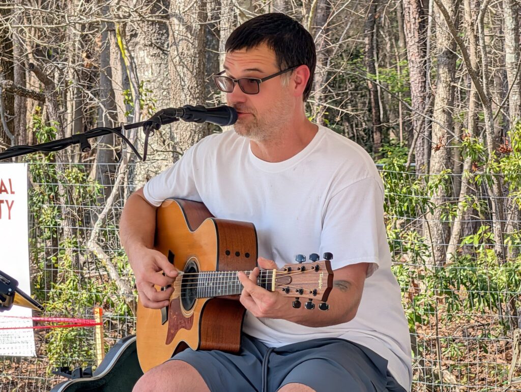 A man wearing sunglasses and a white t-shirt plays an acoustic guitar and sings into a microphone outdoors, with trees in the background.