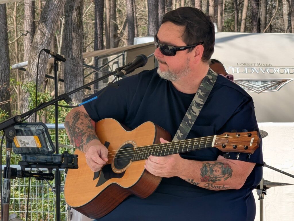 A man wearing sunglasses plays an acoustic guitar outdoors, sitting near a microphone stand, with trees and equipment visible in the background.
