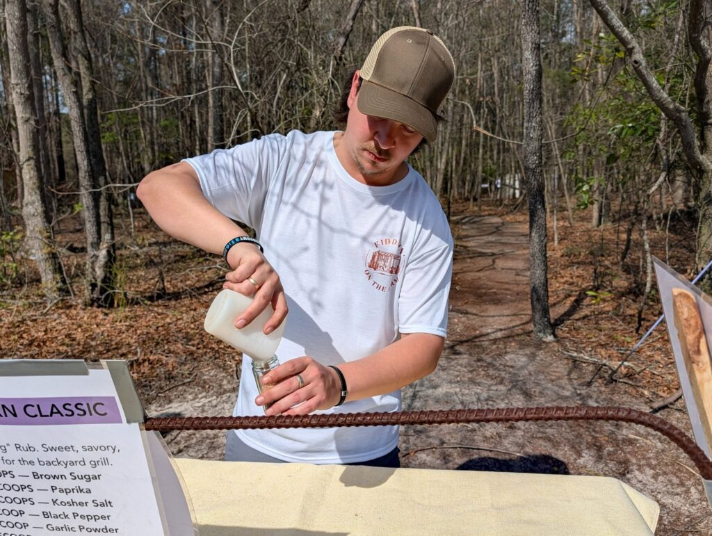 A man in a white shirt and tan cap pours liquid from a bottle into a cup outdoors, with trees and a table in the background.