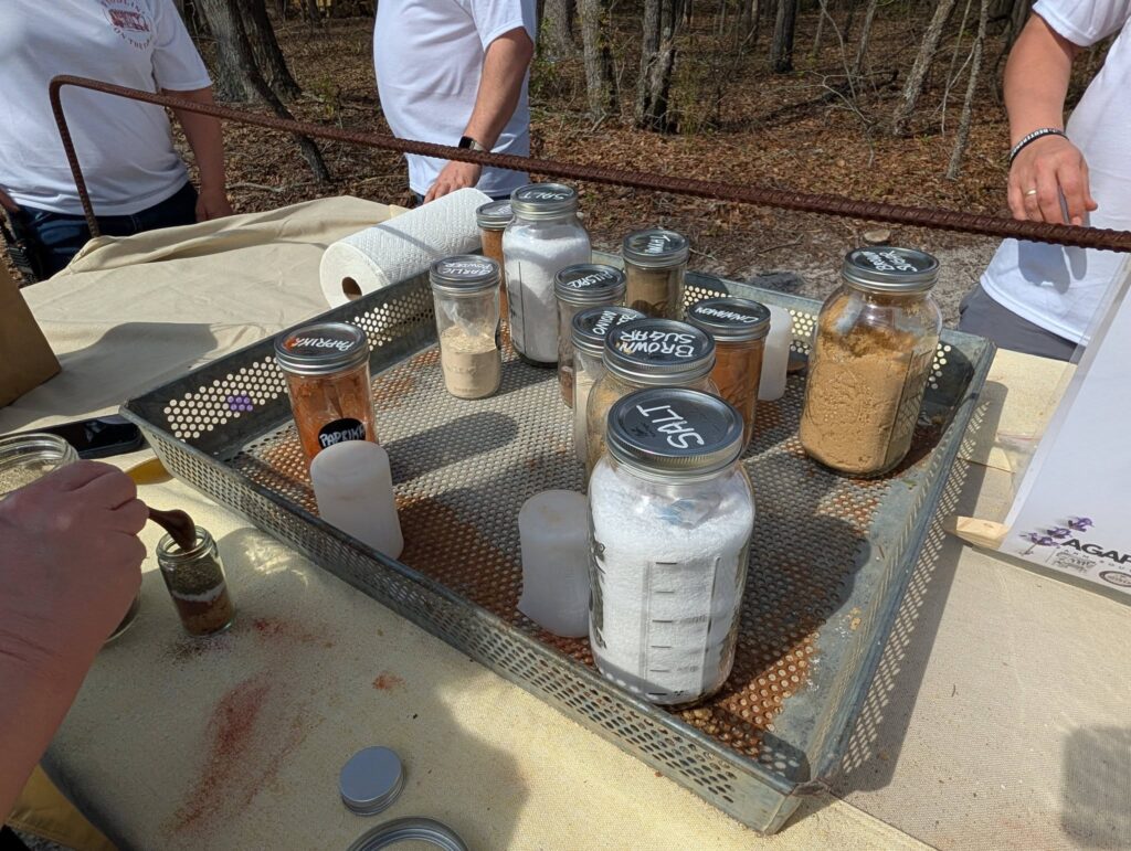 Jars filled with various powders and substances arranged on a mesh tray outdoors, with people standing nearby and trees in the background.