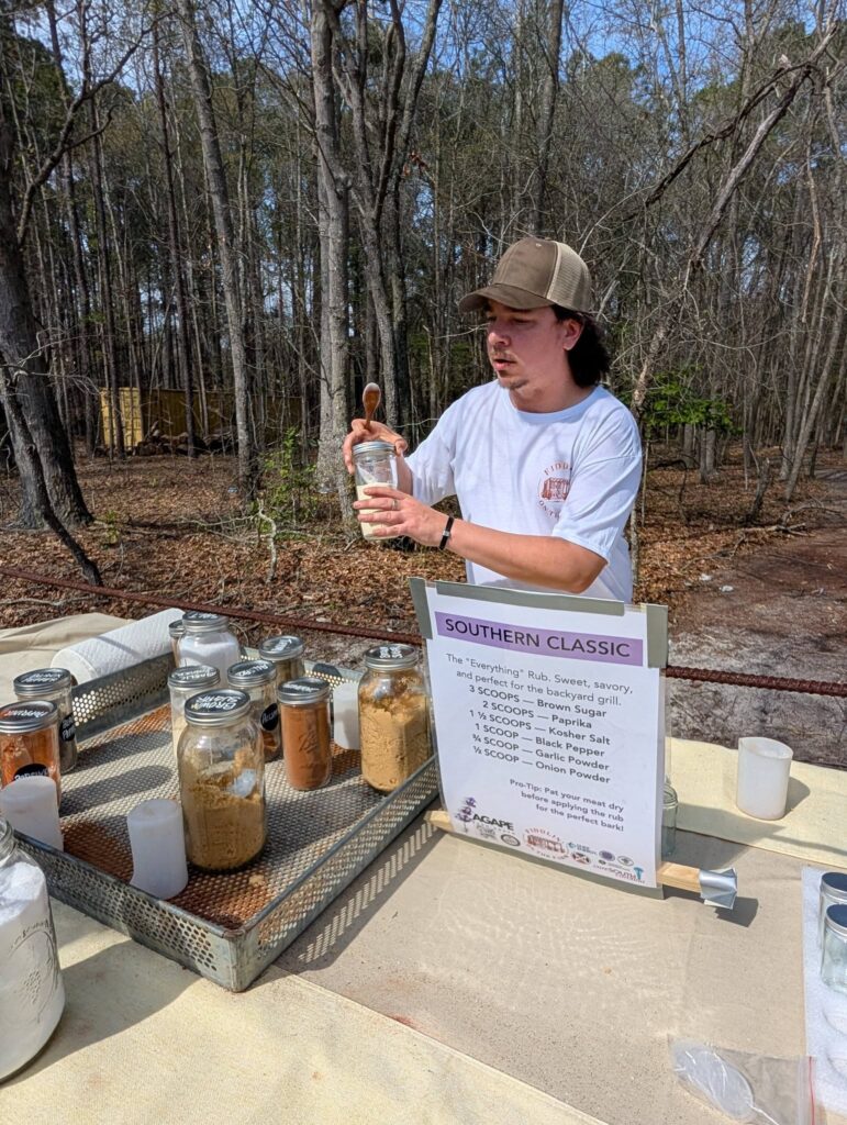 Person outdoors demonstrating with jars of soil samples and a sign, surrounded by trees, on a wooden table with various containers and scientific items.