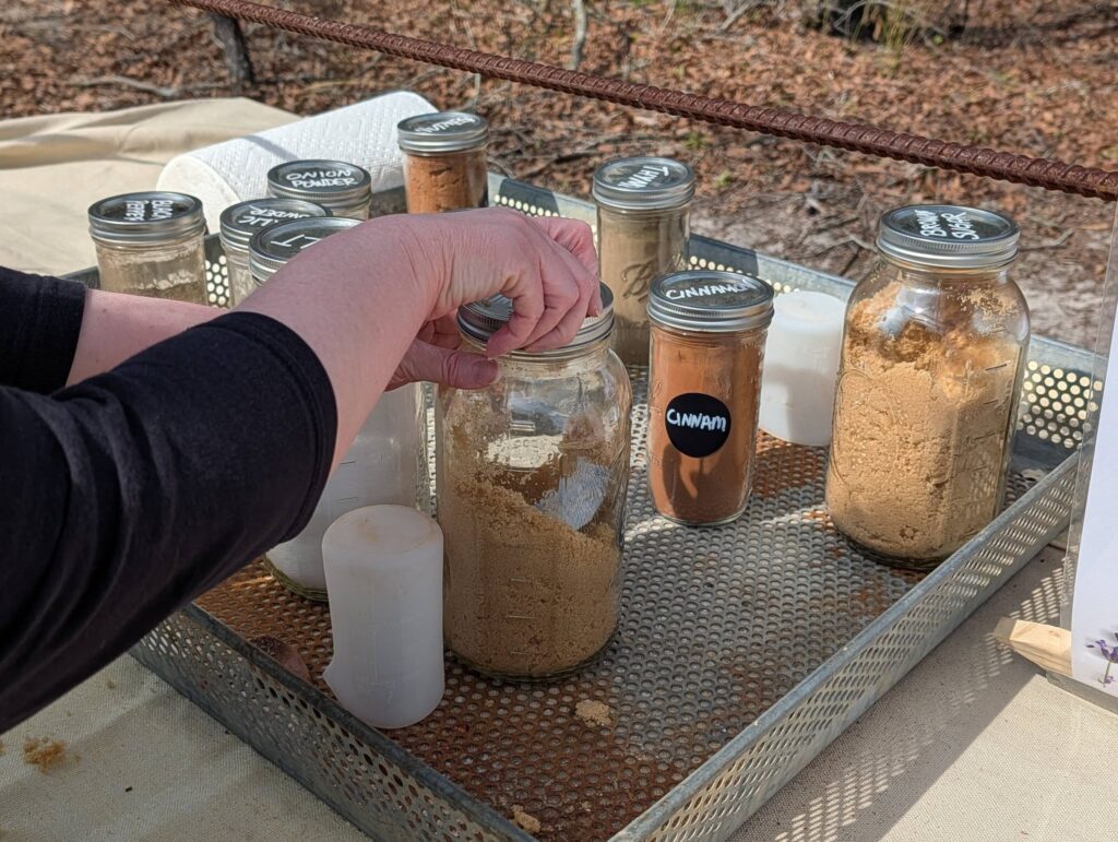 A person’s hand holds a glass jar filled with a tan powder, surrounded by several jars of similar powders on an outdoor table.