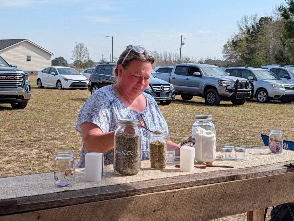 A woman stands outside at a wooden table with large jars of seeds and cups, with parked cars and trees in the background.