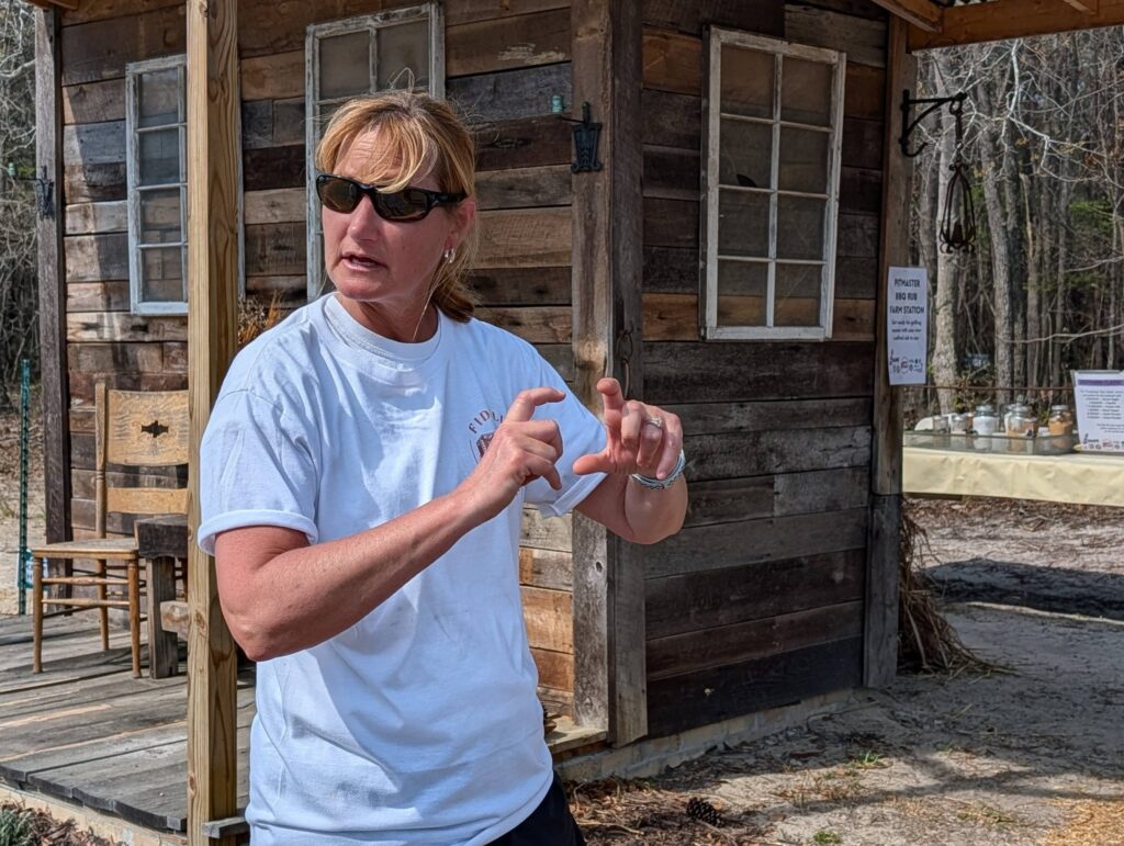 Woman wearing sunglasses and a light shirt gestures with her hands while standing outside in front of a rustic wooden building.