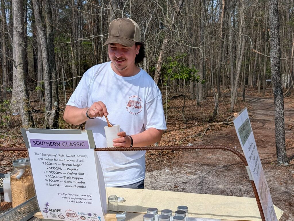 Man wearing a cap and white t-shirt stands behind an outdoor table with jars and informational signs, surrounded by leafless trees.