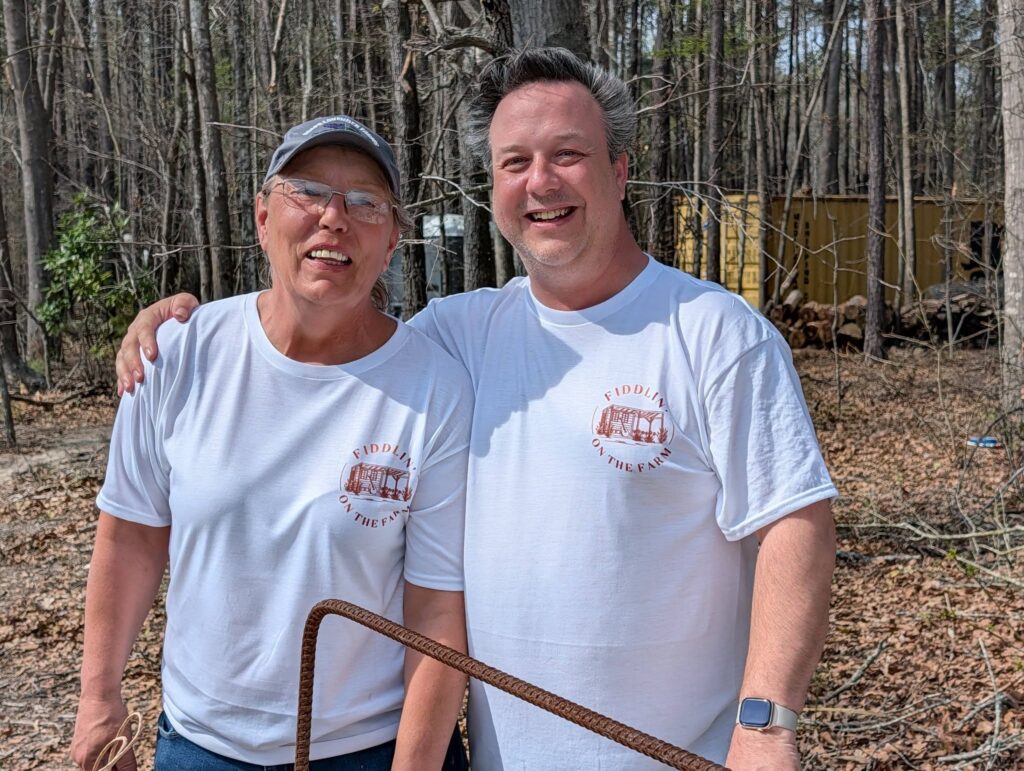Two smiling men in white shirts standing outdoors in a wooded area, one with his arm around the other.