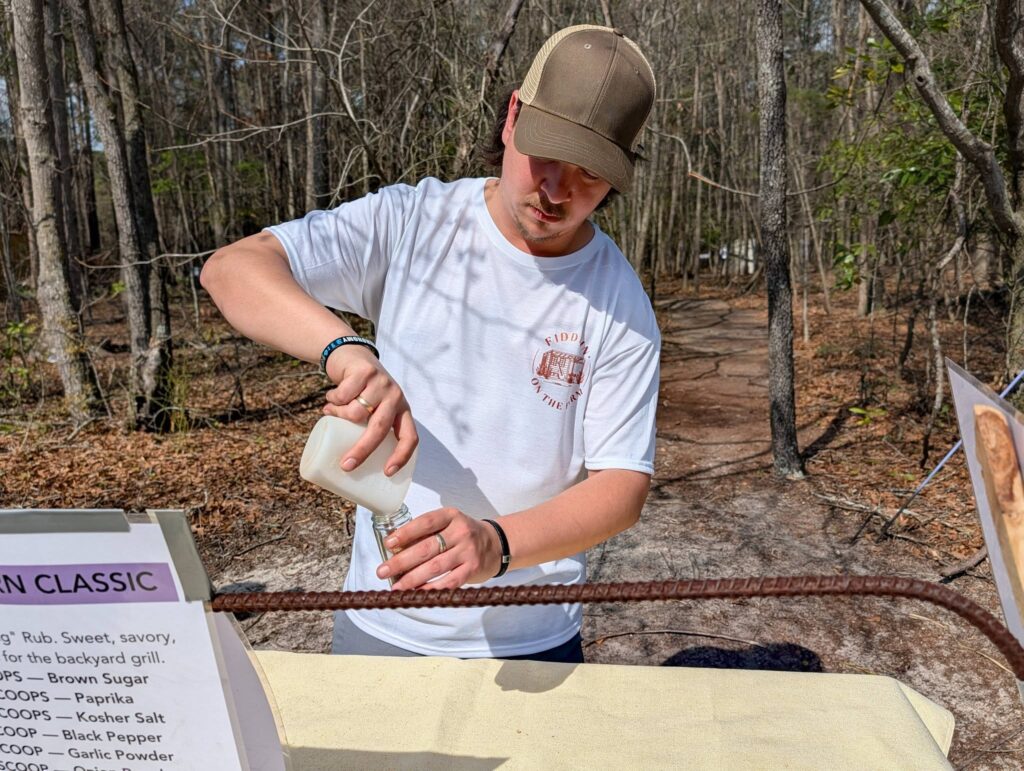 A man outdoors wearing a cap pours a liquid from a bottle into a cup, standing behind a table with informational signage.