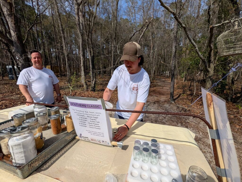 Two people outdoors at a table with jars and eggs, one person reading a sign, surrounded by trees in the background.