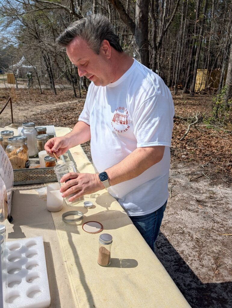 A man in a white shirt works outdoors at a table with various jars and containers, surrounded by trees and natural scenery.