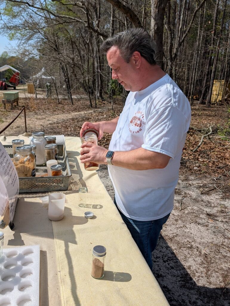A man in a white shirt stands outdoors at a table with various jars and containers, surrounded by trees and a natural setting.