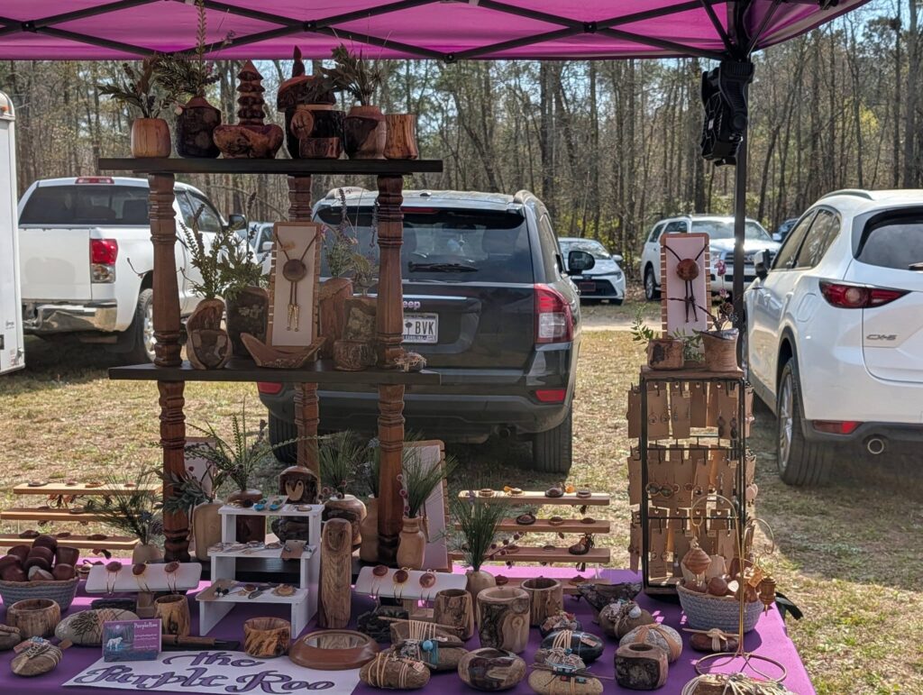 Handcrafted wooden items displayed on a table under a pink canopy at an outdoor market, with parked cars and trees in the background.