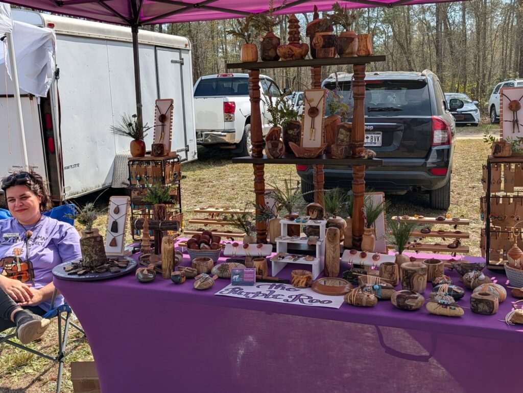 Outdoor market stall with a purple tablecloth displaying various handmade wooden crafts and small decorative items, with cars and trees visible in the background.