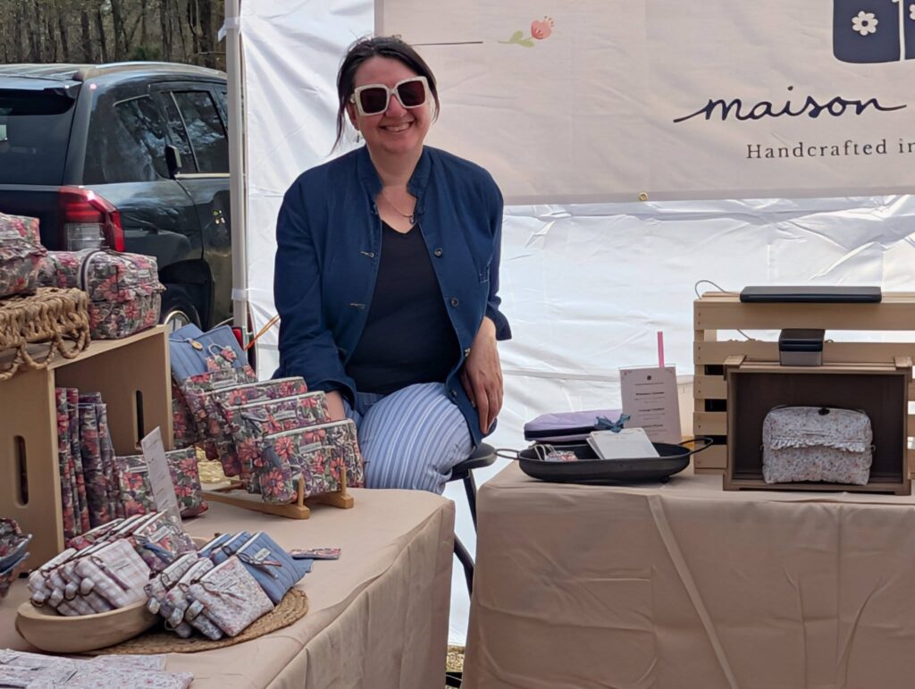 Woman sitting behind a market stall with handmade goods, wearing sunglasses and a blue jacket, smiling at the camera. Products displayed on tables in front of her.