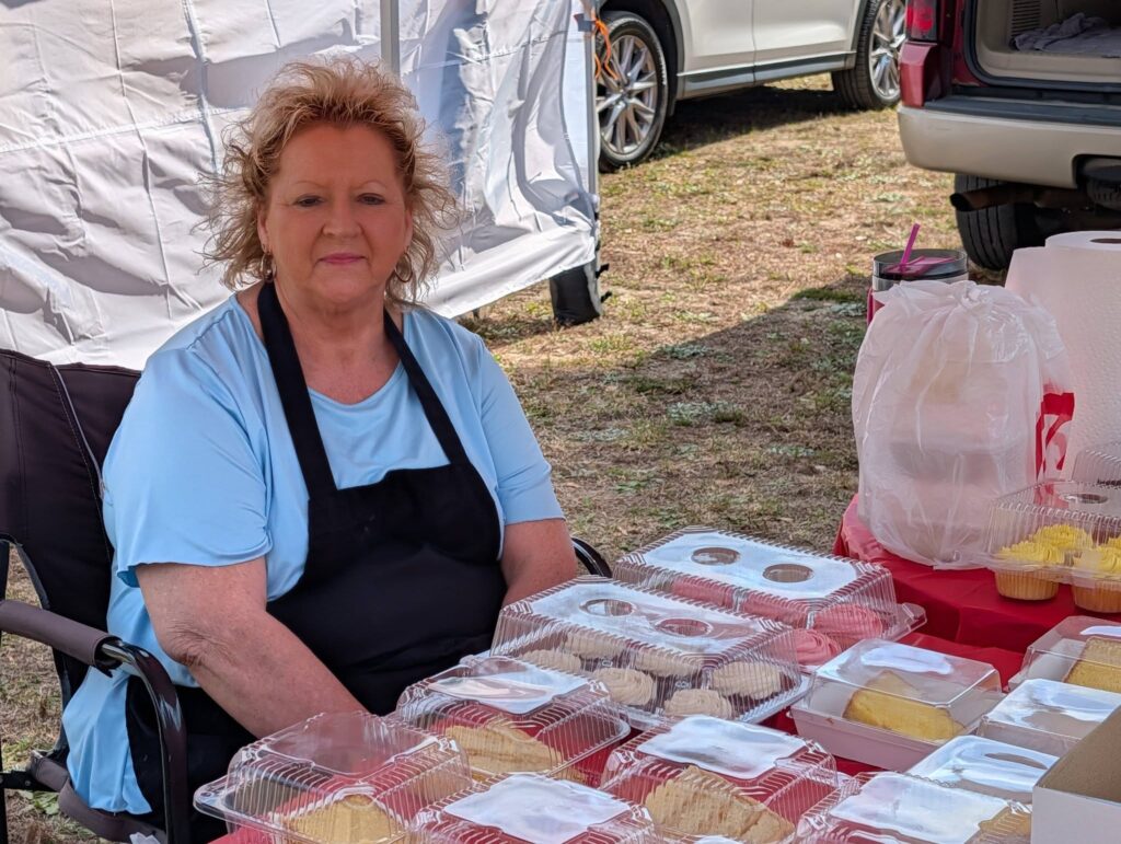 Woman with light hair wearing a blue shirt and black apron sits at an outdoor market table displaying packaged food items.