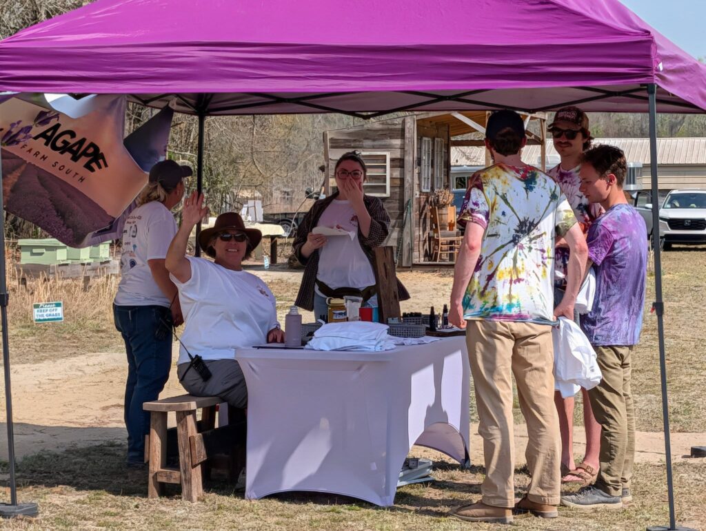 Several people stand and sit under a purple canopy tent at an outdoor event, gathered around a table covered with a white cloth.