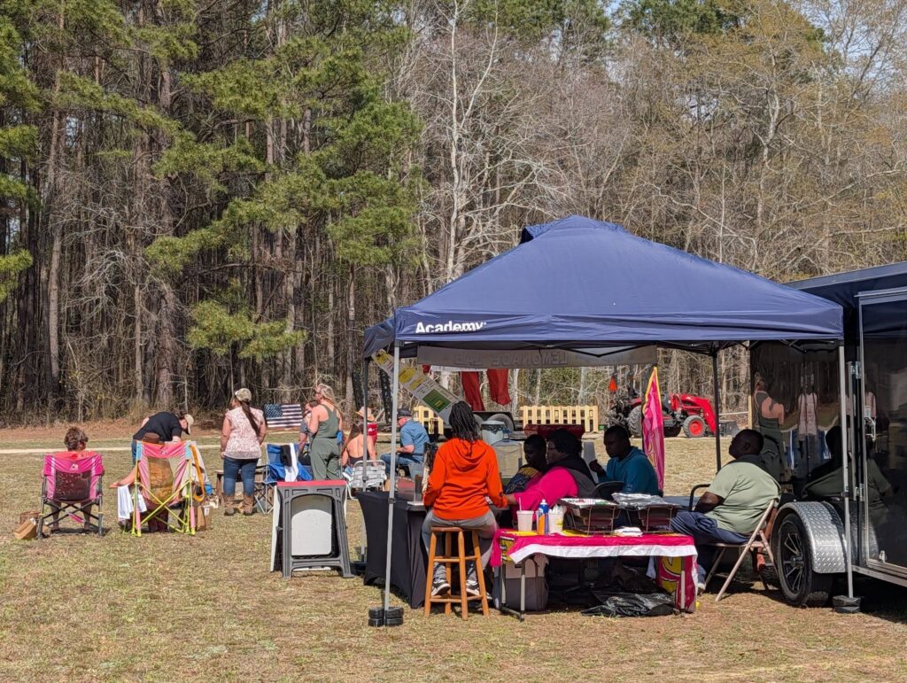 People gathered outdoors under a blue canopy and around tables, enjoying a picnic in a grassy area near trees on a sunny day.