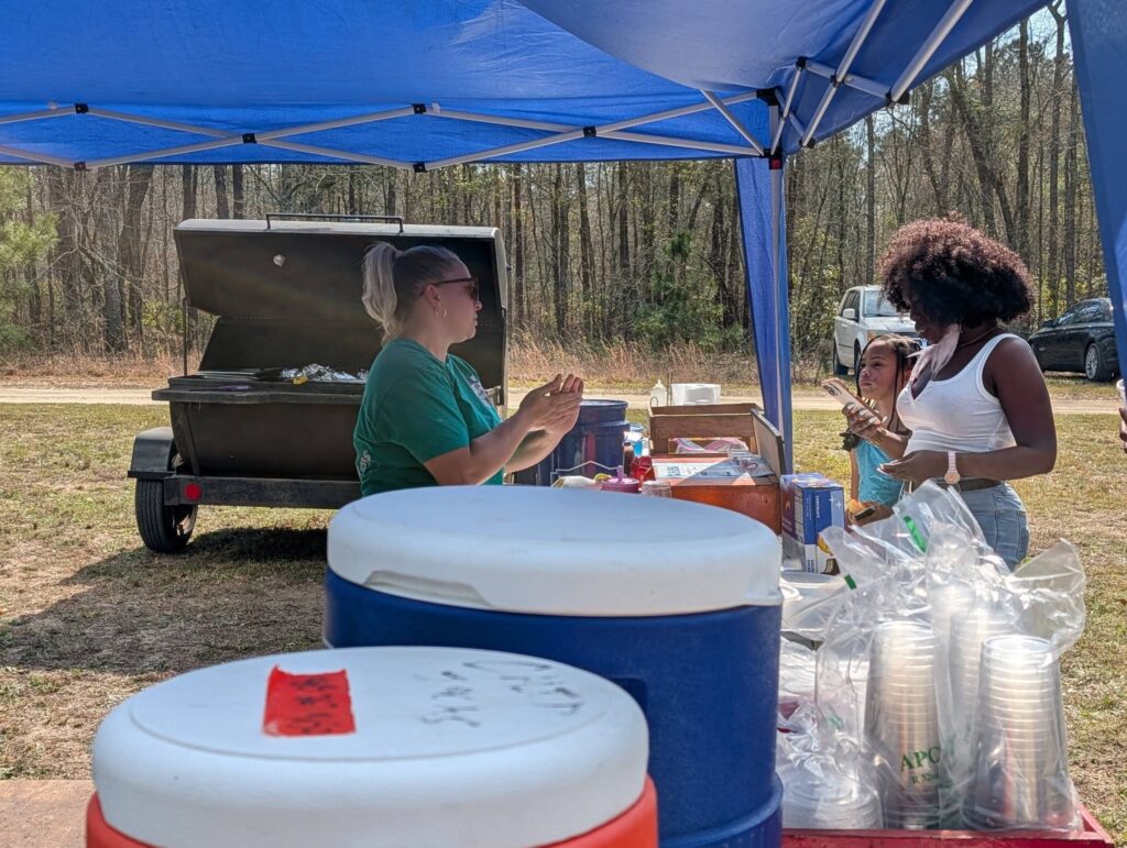 Two people converse under a blue canopy next to coolers and supplies, with a barbecue grill and wooded area in the background.
