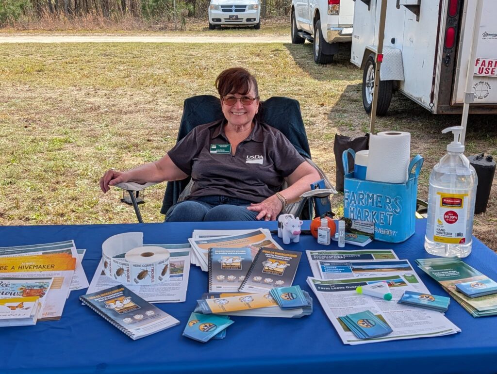 A woman sits behind a table covered with brochures, pamphlets, and informational items at an outdoor event, with vehicles and grass in the background.