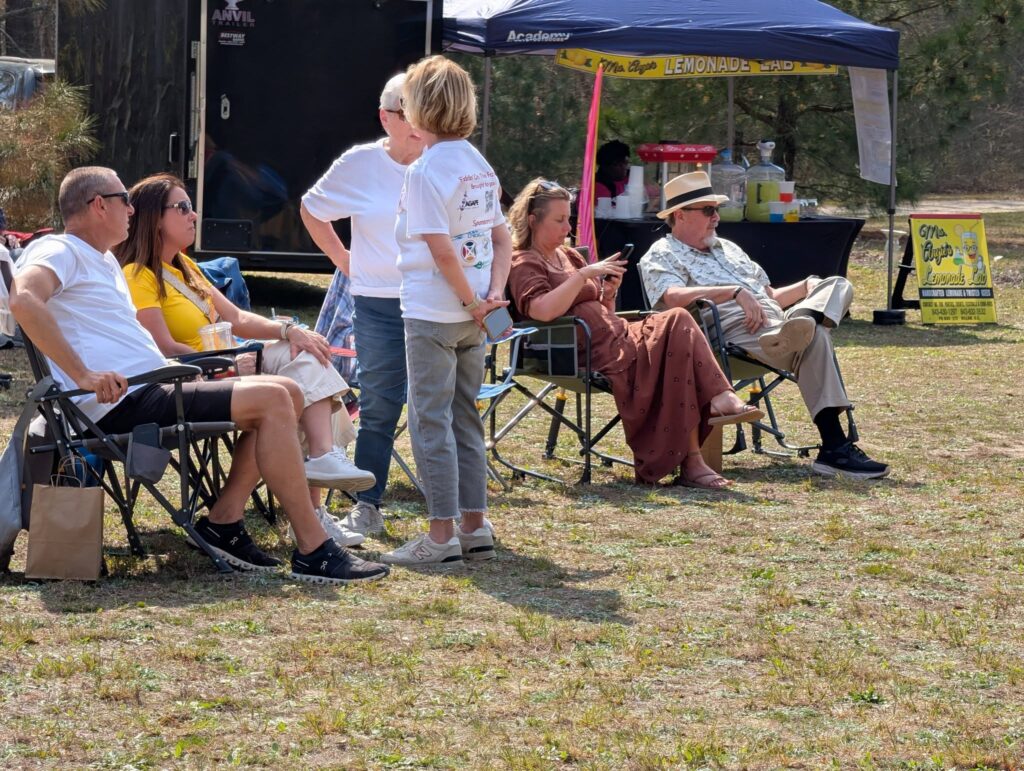 Several people are sitting on folding chairs outdoors, talking and relaxing under canopies on a sunny day. Two women stand and converse while others are seated.
