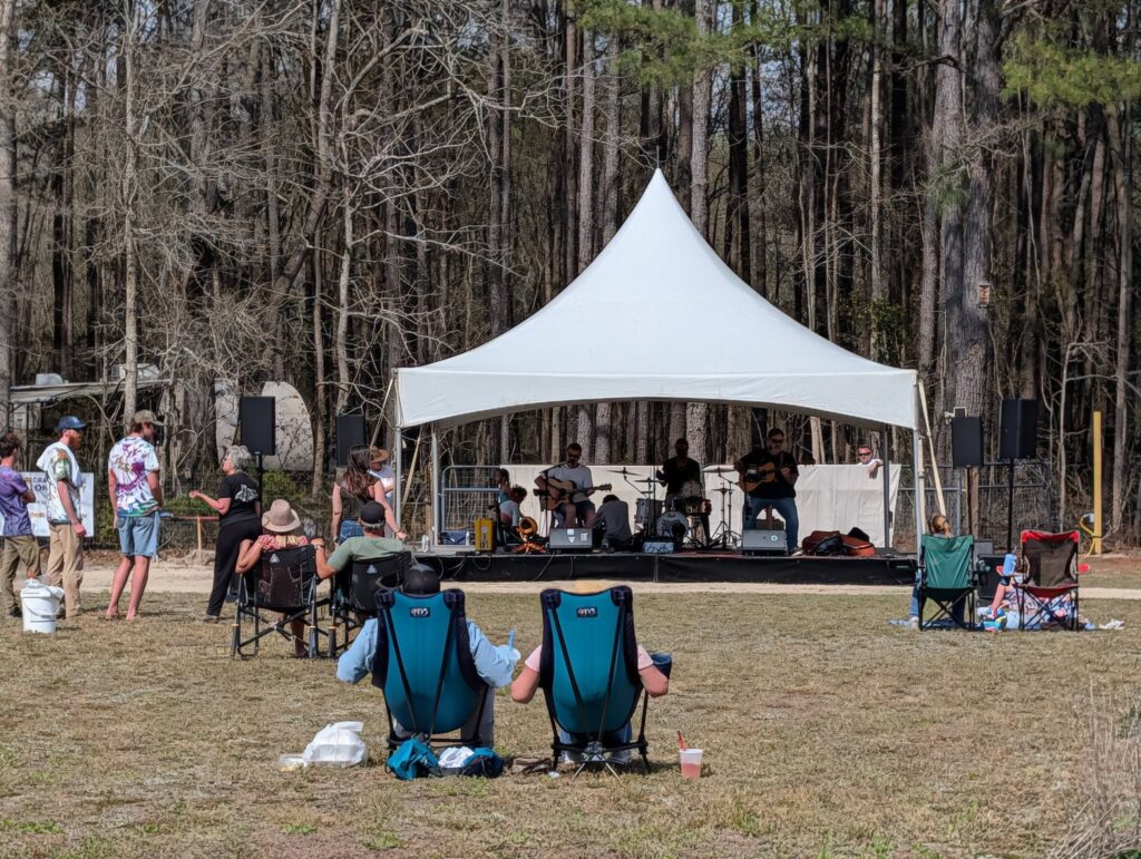 People sitting outside on lawn chairs watching a live band perform on an outdoor stage with a white canopy, surrounded by trees.