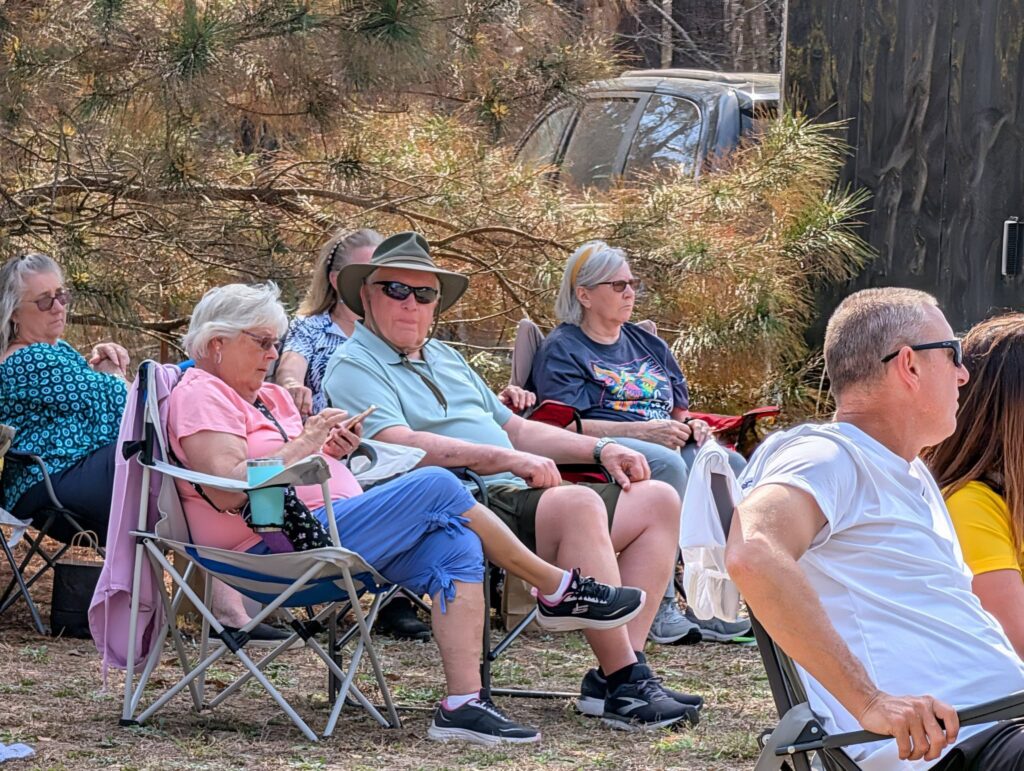Several older adults sitting outdoors on foldable chairs, relaxing and enjoying a sunny day, with trees and a parked car in the background.