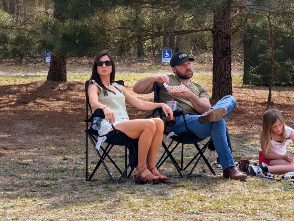 A woman and man sit on folding chairs outdoors in a wooded area, relaxing under trees.