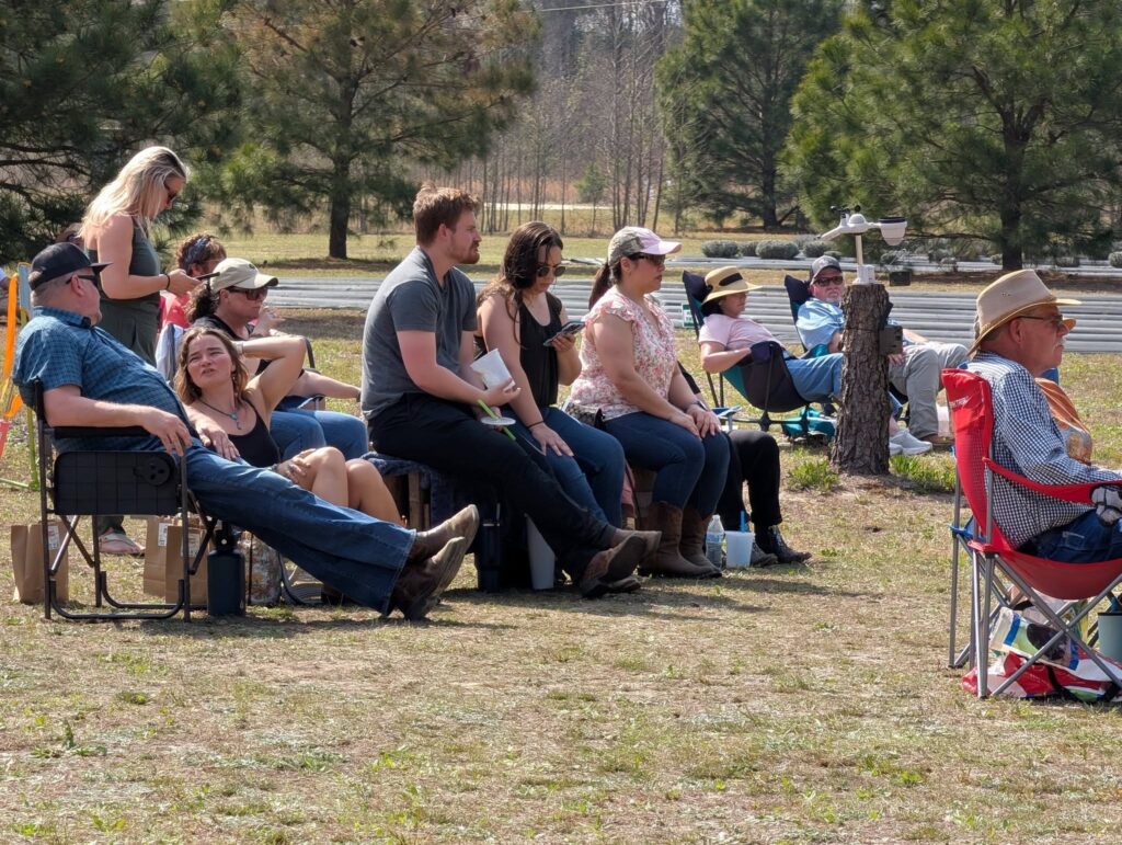 A group of people sitting outdoors on chairs and benches, watching something off-camera on a sunny day, with trees and grass in the background.