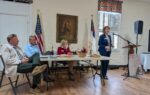 Three people sit at a table with American and Christian flags behind them, while a woman stands at a microphone speaking in a room with wood floors.