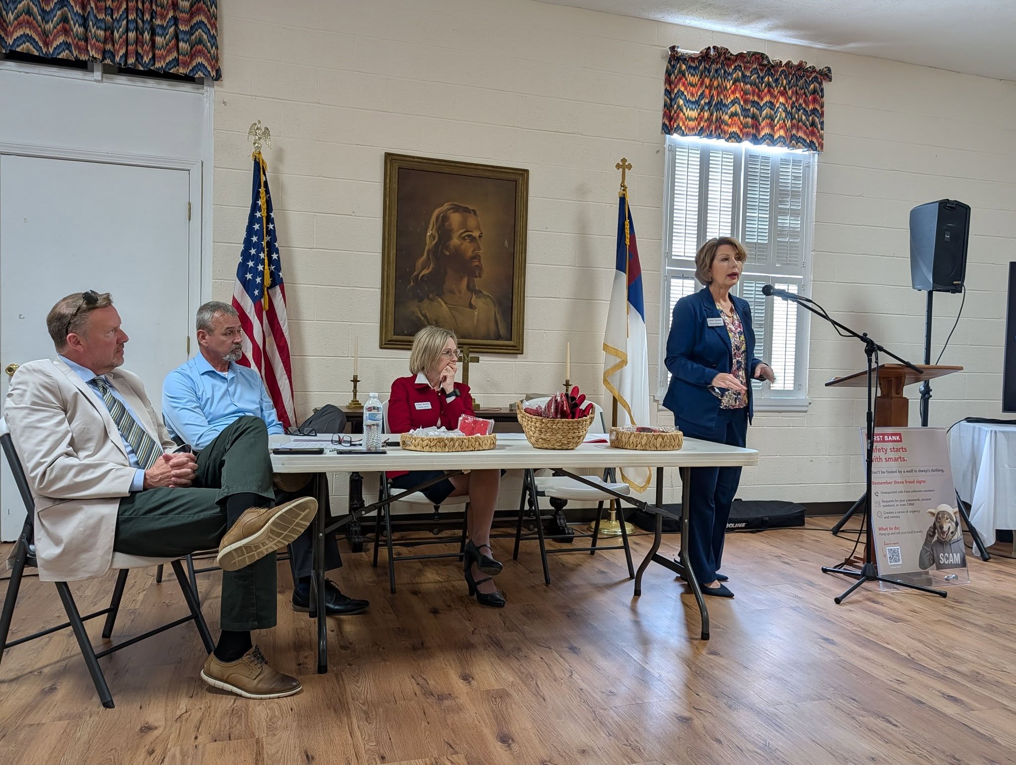 Three people sit at a table with American and Christian flags behind them, while a woman stands at a microphone speaking in a room with wood floors.