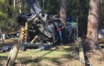 A severely damaged car crashed into trees and a fence, with emergency responders at the scene. Broken utility poles lie on the ground nearby.