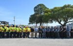 A large group of people, including workers in safety vests and uniforms, stand outside together in a parking lot with trees in the background.