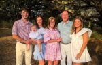 Six people, including a baby, stand together outdoors under large trees, smiling and dressed in light-colored, summery clothes.