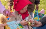 A young child in a red bonnet holds a shiny egg surrounded by adults and toys outdoors on a grassy area.