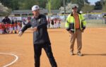 A man in sportswear gestures on a baseball field while another man in a safety jacket stands nearby; children and adults are gathered in the background.