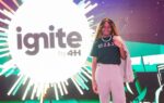 A woman stands smiling in front of a bright sign that reads "Ignite by 4H" with colorful lights in the background.