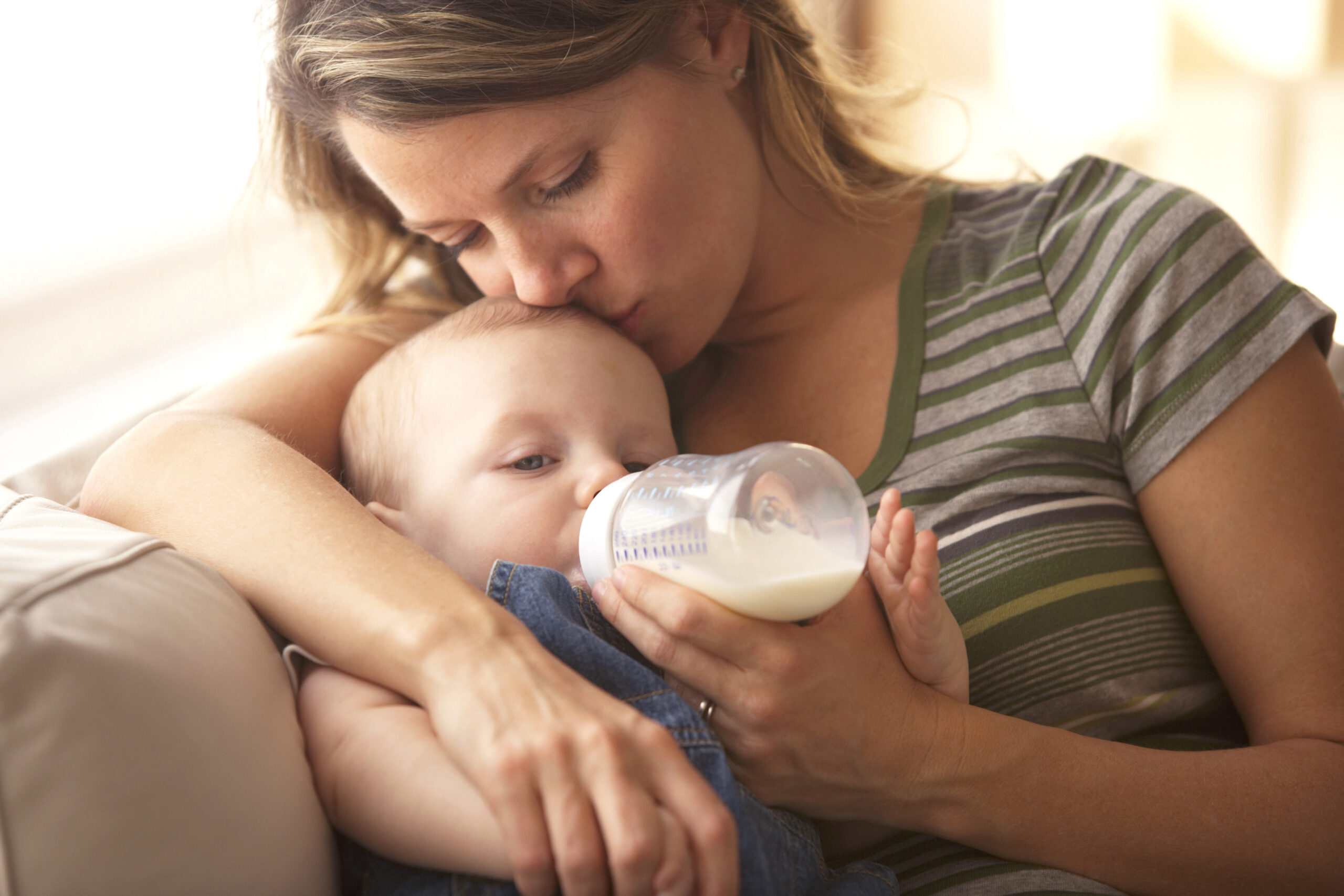 A woman lovingly kisses a baby’s head while feeding the baby with a bottle, sitting together in a cozy setting.