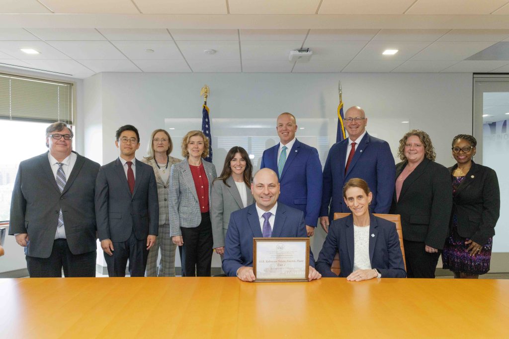 A group of people in business attire pose together, two seated at a table with a plaque, American flags in the background, office setting.