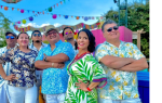A group of people in tropical clothing pose outdoors under colorful decorations. "Furia Tropikal Charlotte N.C." logo is at the top. Bright, sunny weather.