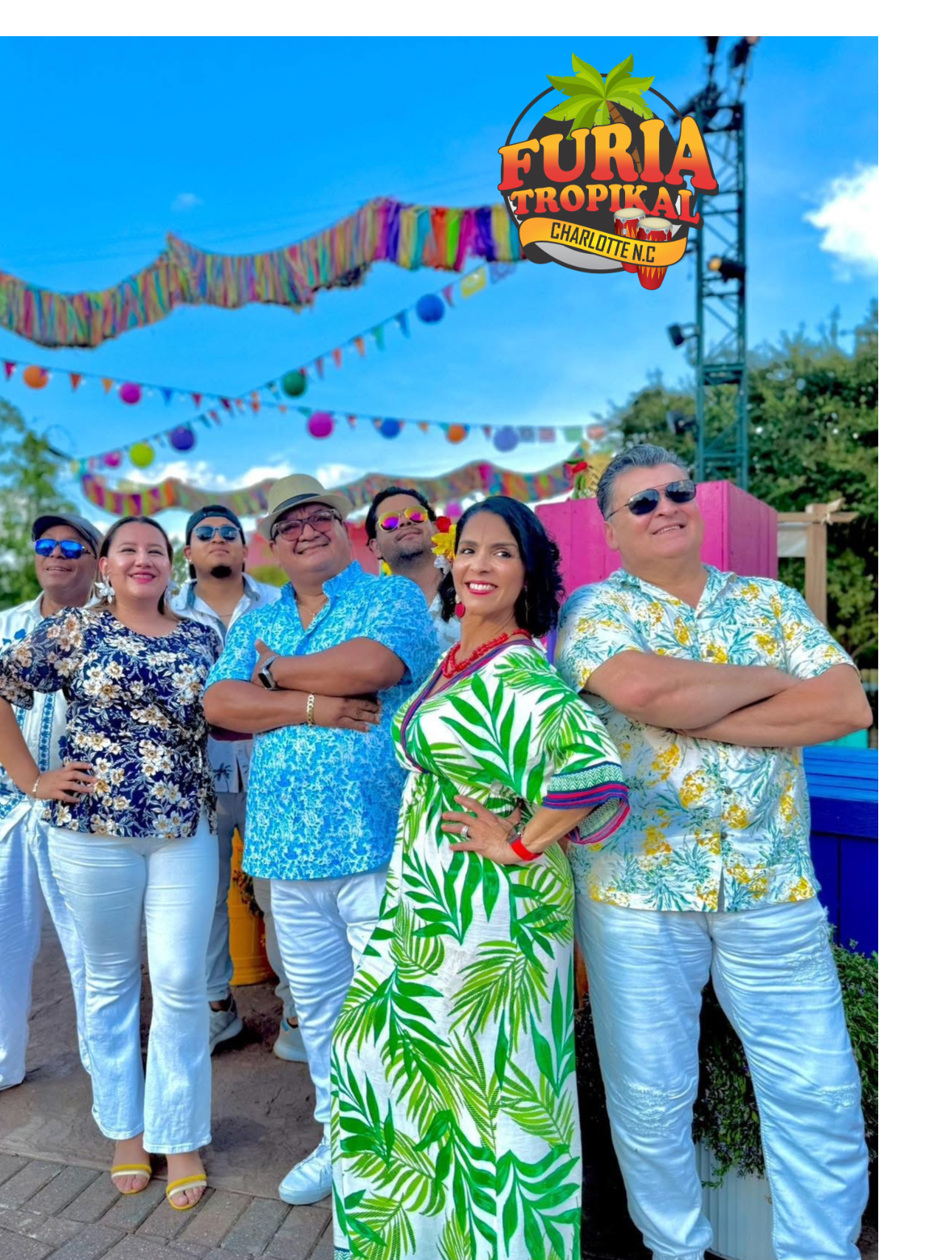 A group of people in tropical clothing pose outdoors under colorful decorations. "Furia Tropikal Charlotte N.C." logo is at the top. Bright, sunny weather.