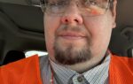 Man wearing safety glasses and an orange safety vest, sitting inside a vehicle.